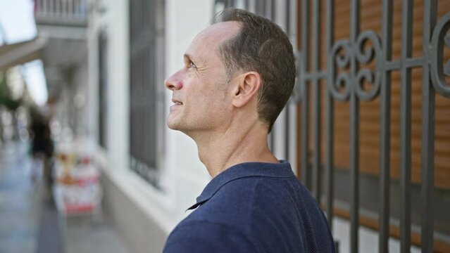 Middle age man, relaxed yet with a serious expression, standing outside looking up to the city's sky from a bustling urban street