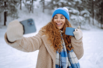Portrait of a beautiful woman in warm clothes takes a selfie with a hot drink from a thermos in her...