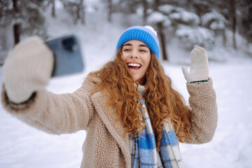 Portrait of a beautiful woman in warm clothes takes a selfie with a hot drink from a thermos in her hands. Happy female traveler with phone enjoying winter day outdoors.