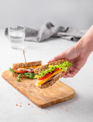 Hands holding a club sandwich with bacon, cheese, tomato and lettuce on a wooden board on a light background.