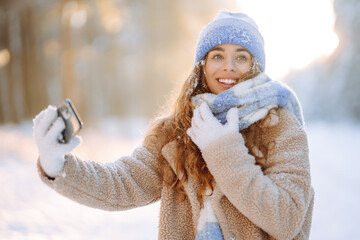 Young woman in a bright hat and scarf with a phone in a snowy forest. Happy female tourist blogs from her phone outdoors. Travel, vacation concept.