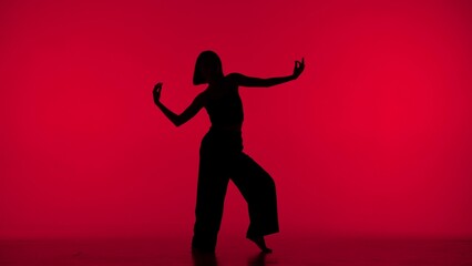 Fototapeta na wymiar In the frame on a red background in the silhouette. Dances slender, beautiful girl. Demonstrates dance moves in the style of hip hop. It is feminine, plastic, rhythmic. General plan