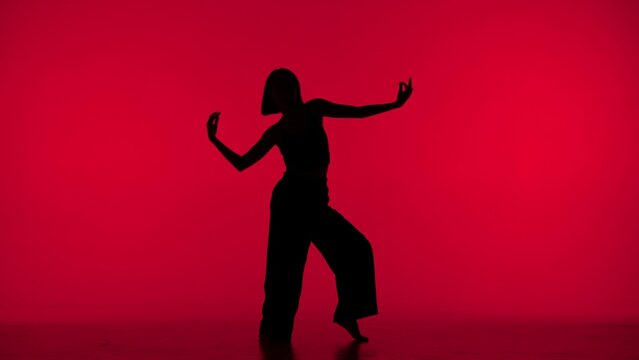 In the frame on a red background in the silhouette. Dances slender, beautiful girl. Demonstrates dance moves in the style of hip hop. It is feminine, plastic, rhythmic. General plan