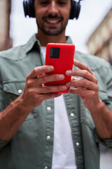 Vertical photo focus on the mobile of a young man using telephone outdoors sending online messages, using wireless headphones to listen music. Copy space.