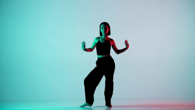 In the frame on a blue, light background. Dances young, beautiful girl. Demonstrates dance moves in the style of hip hop. Shes staring at the camera. Shes feminine in a black top and pants