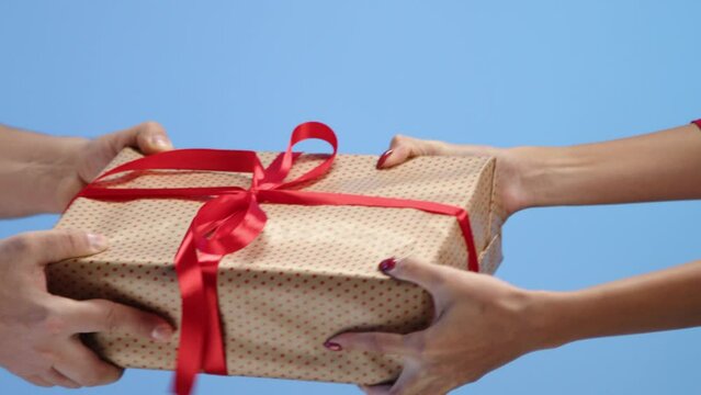 Big box with golden wrapping paper and red thin ribbon with bow on blue background. Male and female hands with red manicure can not share Christmas box. High quality 4k footage