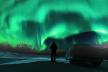 Silhouette of unrecognizable man and dark car on mountain road in front of Northern green lights shine over mountains in Sweden, Lapland. Night photo, Aurora, rear side photo - obrazy, fototapety, plakaty
