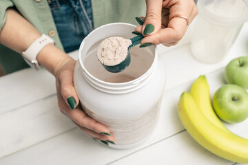 Fototapeta na wymiar Woman in jeans and shirt holding measuring spoon with portion whey protein powder above plastic jar on white wooden table with shaker, banana and apple fruit. Process of making protein drink