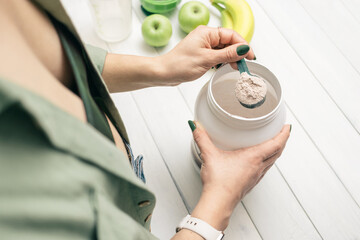 Woman in jeans and shirt holding measuring spoon with portion whey protein powder above plastic jar...