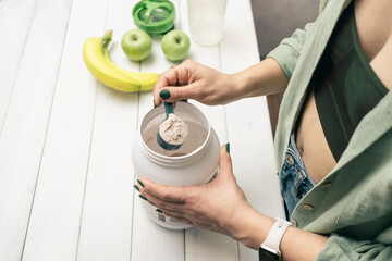 Woman in jeans and shirt holding measuring spoon with portion whey protein powder above plastic jar...