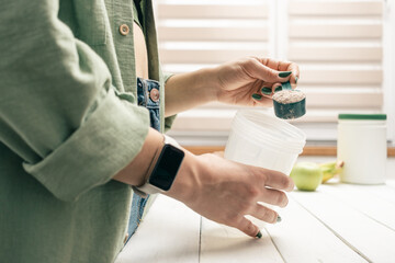 Young woman in jeans and shirt with measuring spoon in her hand puts portion of whey protein powder...