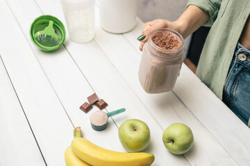 Young woman in jeans and shirt holding glass jar of protein drink cocktail, milkshake or smoothie above white wooden table with measuring spoon of protein powder, chocolate pieces, bananas and apples