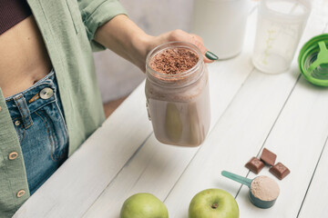 Young woman in jeans and shirt holding glass jar of protein drink cocktail, milkshake or smoothie above white wooden table with measuring spoon of protein powder, chocolate pieces, bananas and apples