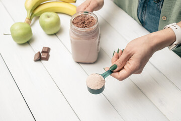 Young woman in jeans and shirt holding measuring spoon with protein powder, glass jar of protein drink cocktail, milkshake or smoothie above white wooden table with chocolate pieces, bananas, apples