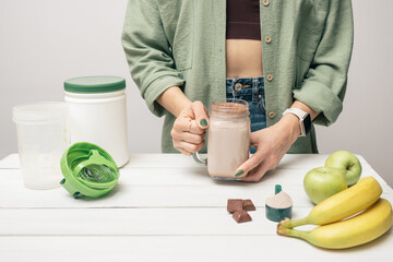 Young woman in jeans and shirt holding glass jar of protein drink cocktail, milkshake or smoothie above white wooden table with measuring spoon of protein powder, chocolate pieces, bananas and apples