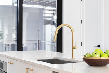 A gold kitchen faucet detail with a white marble countertop and island, a bowl of green apples, and...