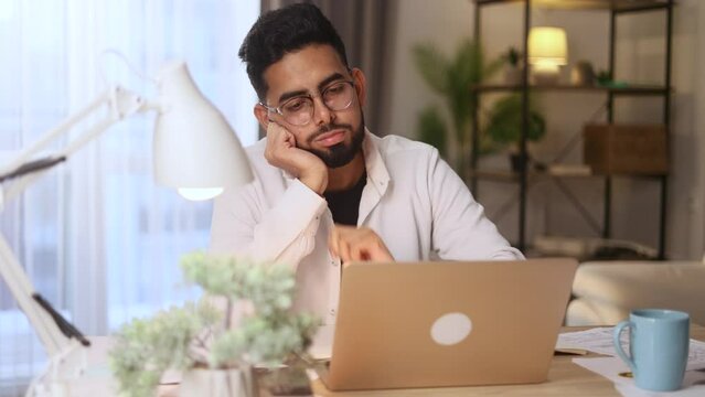 Tired exhausted stressed young man student with glasses preparing for exams and looking at scope of work with sad face at home workplace Overwork education concept
