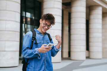 Excited caucasian student guy in jeans shirt holds phone reads message clenches fist smiles wide. Success concept. Teenager celebrating win, passed exam. Happy people. - obrazy, fototapety, plakaty