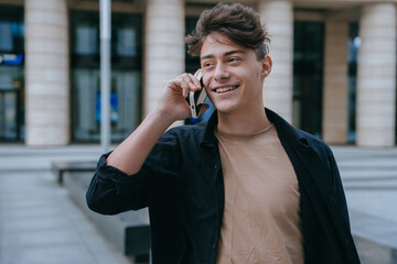 Happy teenager boy talks by phone outside looks aside broad smiling. Handsome caucasian boy in love...