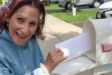 Happy older senior citizen woman smiles as she removes a blank white envelope from a white mailbox
