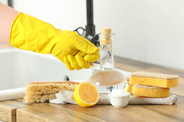 Female hand in rubber glove with jug of vinegar, sponges, brush, baking soda and lemon on wooden...