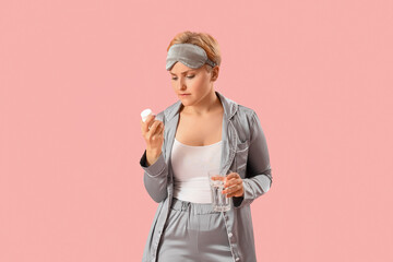 Young woman in pajamas with pill bottle and glass of water on pink background