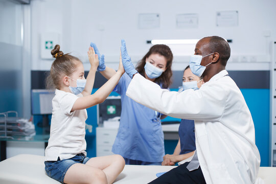 At a checkup in an office, a doctor and nurse give a little youngster and her mother a high five. Image showcasing a friendly environment between medical staff and patients. Face masks for protection.