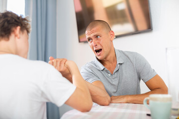 Young boy competing with his father in arm wrestling on kitchen table. - obrazy, fototapety, plakaty