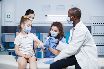 Multiracial doctor and nurse in consultation room caring for young patient and her mother. Male physician and female medical assistant wrapping sterilized bandage on the arm of a little girl.