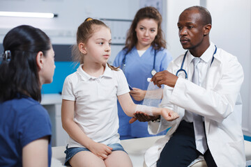 Fototapeta premium Arm of girl patient being meticulously wrapped with a sterile bandage by multicultural medical personnel. Caucasian youngster accompanied by her mother is being treated by doctor and nurse.
