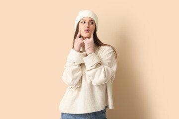 Young woman in winter clothes blowing kiss on beige background