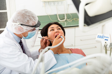 Dentist in face mask treating asian female patient using dental drill in dentist office