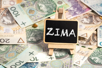 a small wooden writing board standing on scattered Polish zloty PLN banknotes, a chalk inscription "Zima" on the black board. translation: winter (selective focus)