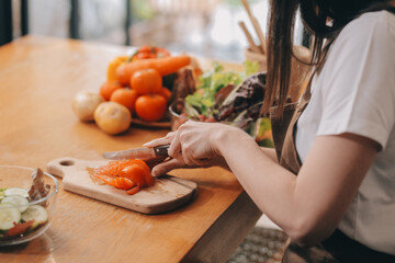 Delicious fruit and vegetables on a table and woman cooking. Housewife is cutting green cucumbers on a wooden board for making fresh salad in the kitchen.