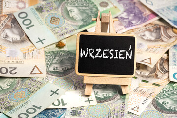 a small wooden writing board standing on scattered Polish zloty PLN banknotes, a chalk inscription "Wrzesień" on the black board. translation: July (selective focus)