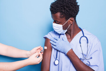 Caucasian person with syringe administers vaccinations to African American medical assistant. Medic using vaccine injection on black male nurse on arm for health and immunity. - obrazy, fototapety, plakaty