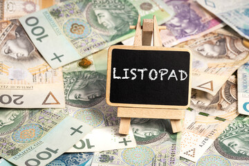 a small wooden writing board standing on scattered Polish zloty PLN banknotes, a chalk inscription "Listopad" on the black board. translation: November (selective focus)