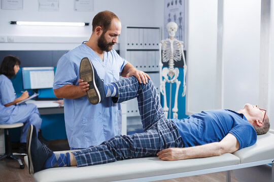 Physiotherapist performs leg exercise to stretch muscles and crack bones of elderly man in hospital cabinet. Retired patient receives kinesiology treatment for knee injury with assistance from nurse.