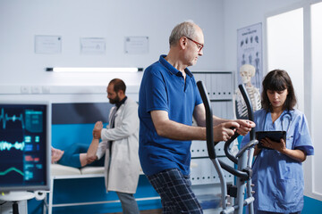 Elderly male patient is engaged in physiotherapy on stationary bicycle, with support from nurse in blue scrubs. Medical assistant with digital tablet helping senior man with physical therapy. - Powered by Adobe