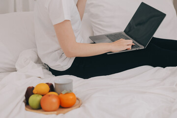 Efficient Young Millennial Girl Sitting on a Bed in the Morning, Uses Laptop Computer and Eats Croissants and Drinks Coffee for Breakfast