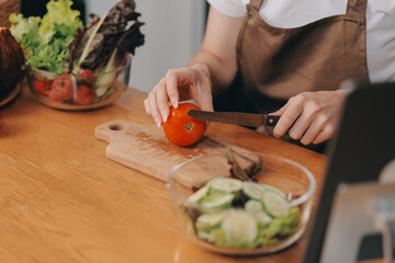 Delicious fruit and vegetables on a table and woman cooking. Housewife is cutting green cucumbers on a wooden board for making fresh salad in the kitchen.