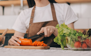 Delicious fruit and vegetables on a table and woman cooking. Housewife is cutting green cucumbers on a wooden board for making fresh salad in the kitchen.