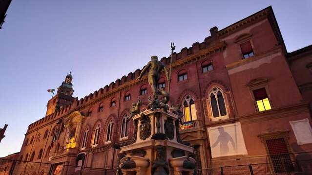Under the night's cloak, the Neptune statue in Bologna stands majestically, casting an imposing silhouette against the backdrop of the illuminated Accursio Palace with Italian flag.