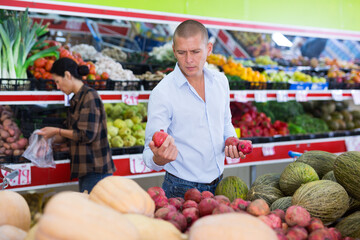 Interested adult man shopping at farmers market, choosing ripe pomegranates in fruit and vegetable department..