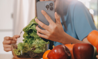 A young woman with a beautiful face in a blue shirt with long hair eating fruit sitting inside the kitchen at home with a laptop and notebook for relaxation, Concept Vacation.