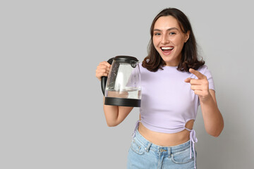 Happy young woman with modern electric kettle pointing at viewer on grey background