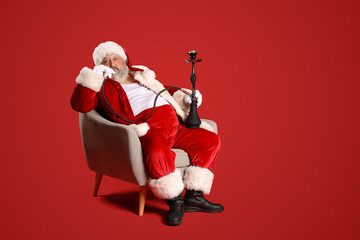 Bad Santa Claus with hookah sitting in armchair on red background - obrazy, fototapety, plakaty