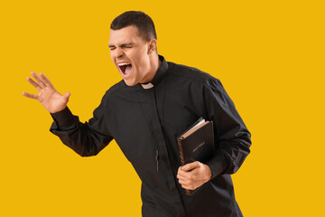 Young priest with Holy Bible shouting on yellow background - obrazy, fototapety, plakaty