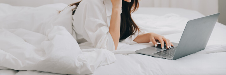 Happy young female in casual clothes smiling and looking at camera while using laptop on bed in bedroom at home