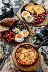 Set of delicious breakfasts, fried eggs with toasts and sausage on hot pan, english breakfast with beans and omelette with cheese on pan over wooden background. Healthy food, top view